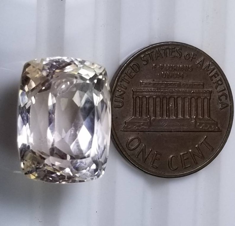 May include: A faceted, light-colored gemstone with a rectangular cushion cut, next to a United States one-cent coin. The gemstone has a translucent appearance and reflects light, showcasing its multiple facets. The coin is bronze-colored and displays the words "United States of America."