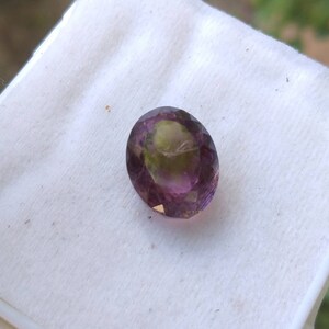 May include: An oval-cut gemstone with a blend of colors, including purple, green, and hints of yellow. The gem is set against a white background, showcasing its facets and internal color variations. The stone's polished surface reflects light, highlighting its clarity.
