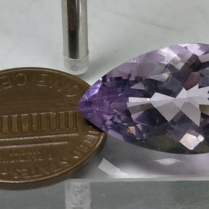 Puede incluir: Una piedra preciosa de amatista lavanda facetada en forma de pera. La gema est&aacute; junto a un centavo estadounidense para comparar el tama&ntilde;o. Una herramienta plateada est&aacute; en el fondo. La piedra preciosa tiene una superficie pulida y facetas internas.