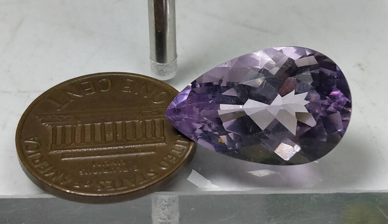 Puede incluir: Una piedra preciosa de amatista lavanda facetada en forma de pera. La gema est&aacute; junto a un centavo estadounidense para comparar el tama&ntilde;o. Una herramienta plateada est&aacute; en el fondo. La piedra preciosa tiene una superficie pulida y facetas internas.