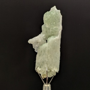 Op de afbeelding: Een lichtgroen mineraal specimen met een kristallijne structuur, gemonteerd op een zilveren metalen standaard. Het mineraal heeft een ruw, getextureerd oppervlak en een doorschijnend uiterlijk. De achtergrond is effen zwart.