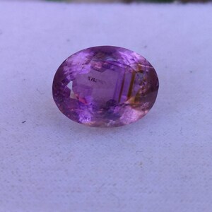 May include: An oval-cut gemstone with a gradient of colors, primarily purple and amber. The gem is set against a white background, showcasing its polished surface and internal facets. This gemstone is suitable for jewelry making.