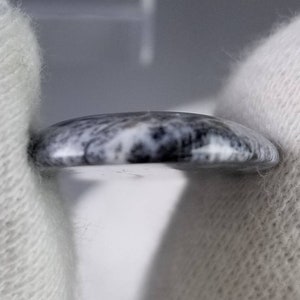 Pode incluir: Uma pequena pedra redonda e polida com um padr&atilde;o marmorizado cinza e branco. A pedra &eacute; segurada entre duas m&atilde;os enluvadas brancas, mostrando sua superf&iacute;cie lisa e curva. O design da pedra lembra pedra natural.
