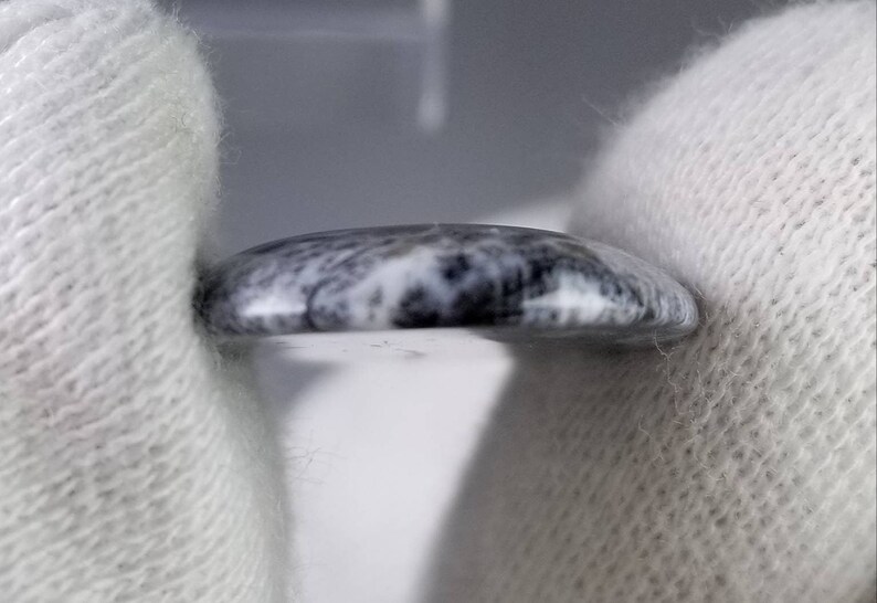 Pode incluir: Uma pequena pedra redonda e polida com um padr&atilde;o marmorizado cinza e branco. A pedra &eacute; segurada entre duas m&atilde;os enluvadas brancas, mostrando sua superf&iacute;cie lisa e curva. O design da pedra lembra pedra natural.