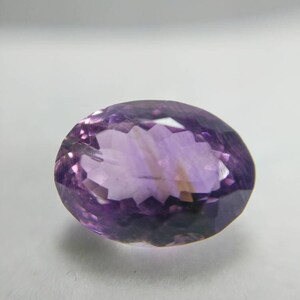 May include: An oval-cut amethyst gemstone. The gem is a deep purple color with facets that catch the light. The gemstone is set against a white background.