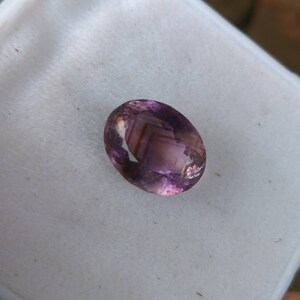 May include: An oval-cut gemstone with a blend of purple and pink hues. The gem has internal striations and facets that catch the light, creating a shimmering effect. The stone is set against a white background.