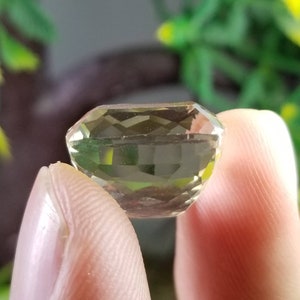 May include: A faceted, light green gemstone held between fingers. The gem has a rectangular shape with multiple facets, reflecting light. The background is blurred, with green and yellow foliage.