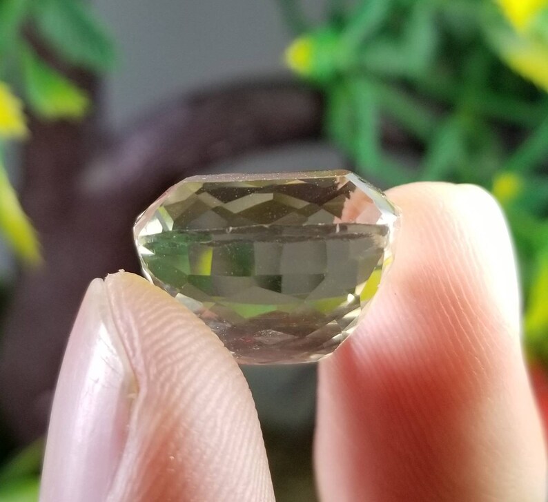 May include: A faceted, light green gemstone held between fingers. The gem has a rectangular shape with multiple facets, reflecting light. The background is blurred, with green and yellow foliage.