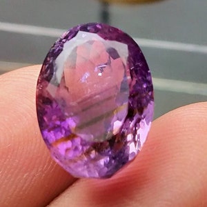 Könnte beinhalten: Ein facettierter, ovaler Amethyst-Edelstein. Der Edelstein zeigt einen Farbverlauf von Lilatönen, mit helleren Schattierungen in der Mitte und dunkleren Tönen zu den Rändern hin. Der Schliff des Steins erzeugt mehrere reflektierende Oberflächen.