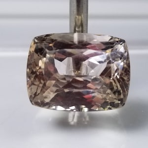 May include: A faceted, cushion-cut gemstone with a light brown hue. The gem is held by a silver metal rod, showcasing its intricate internal structure and reflective surfaces. The gemstone's shape is square with rounded corners.