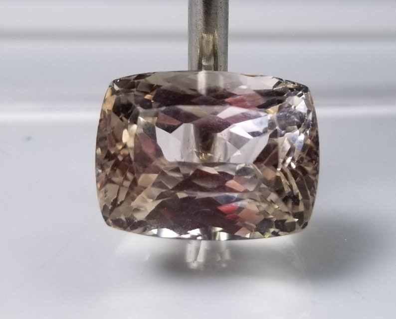 May include: A faceted, cushion-cut gemstone with a light brown hue. The gem is held by a silver metal rod, showcasing its intricate internal structure and reflective surfaces. The gemstone's shape is square with rounded corners.
