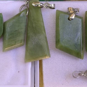 Pode incluir: Uma coleção de pingentes de jade verde em várias formas geométricas, incluindo retângulos, losangos e trapézios. Cada pingente tem uma argola prateada para prender a uma corrente. O jade tem uma superfície lisa e polida.