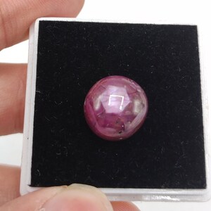 May include: A polished, round ruby gemstone, presented on a black velvet surface within a white box. The ruby is a deep red colour with subtle internal inclusions. The gemstone is held by a person's hand.