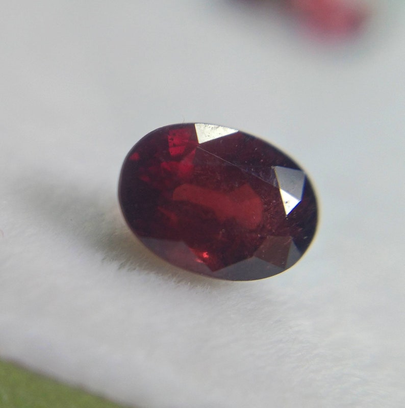 May include: A deep red, oval-cut gemstone with multiple facets. The polished surface reflects light, creating a sparkling effect. The gem is set against a white background. The stone's dimensions are not visible.