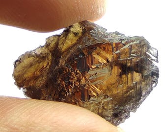 May include: A rough, uncut gemstone with a mix of brown and amber hues. The mineral has a crystalline structure with visible facets and a slightly translucent appearance. The stone is held between two fingers against a white background.