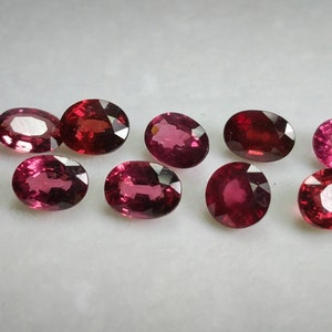 May include: A collection of oval gemstones in shades of red and pink. The polished gems are displayed on a white surface, highlighting their facets and colours. These gems are likely for jewellery making, with each stone having a unique hue.