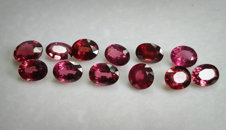 May include: A collection of oval gemstones in shades of red and pink. The polished gems are displayed on a white surface, highlighting their facets and colours. These gems are likely for jewellery making, with each stone having a unique hue.