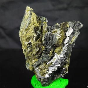 May include: A close-up of a mineral specimen featuring a cluster of dark, metallic-looking crystals alongside a lighter, olive-green crystalline structure. The mineral sits on a bright green base against a dark background.