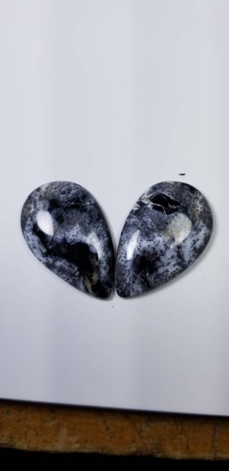 Pode incluir: Duas pedras preciosas em forma de gota com um padr&atilde;o marcante de redemoinhos e manchas pretas e brancas. As pedras polidas t&ecirc;m um acabamento brilhante e est&atilde;o sobre um fundo branco.
