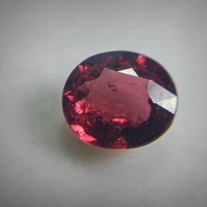 May include: A round-cut, faceted gemstone in a deep red colour. The gem has a polished surface with multiple facets that reflect light, creating a sparkling effect. The gemstone is set against a plain, light background.