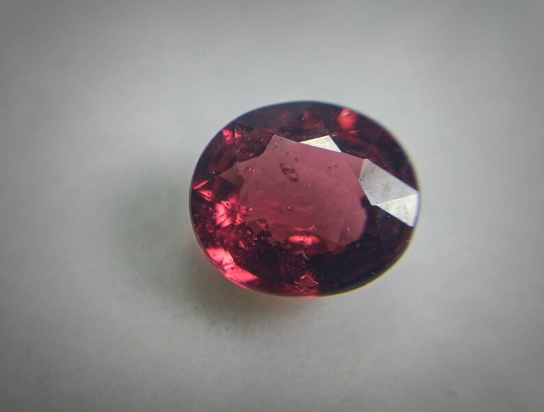 May include: A round-cut, faceted gemstone in a deep red colour. The gem has a polished surface with multiple facets that reflect light, creating a sparkling effect. The gemstone is set against a plain, light background.