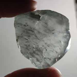 Puede incluir: Una gema translúcida de color azul pálido, con inclusiones internas y una forma irregular. La piedra se sostiene entre los dedos, mostrando su textura natural y sutiles variaciones de color. La gema parece ser un espécimen en bruto, sin cortar.