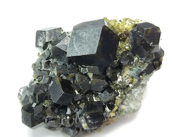 May include: A cluster of dark, cubic crystals, possibly a mineral specimen, with a deep, almost black color. The crystals are densely packed, with some lighter green and yellow accents. The overall structure is rough and textured, set against a white background.