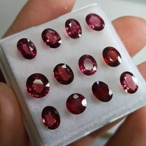 May include: A selection of oval-shaped, deep red gemstones presented in a white tray. The gems have a polished surface, reflecting light and revealing their internal colour. The arrangement showcases the different sizes and shades within the collection.