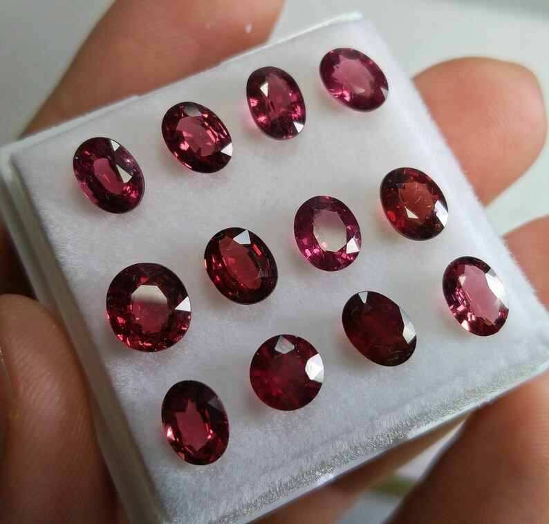 May include: A selection of oval-shaped, deep red gemstones presented in a white tray. The gems have a polished surface, reflecting light and revealing their internal colour. The arrangement showcases the different sizes and shades within the collection.