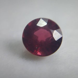 May include: A round, faceted, deep red gemstone. The gem has a polished surface with multiple facets that reflect light. The stone is set against a plain, light background, highlighting its colour and shape. This gemstone could be used in jewellery.