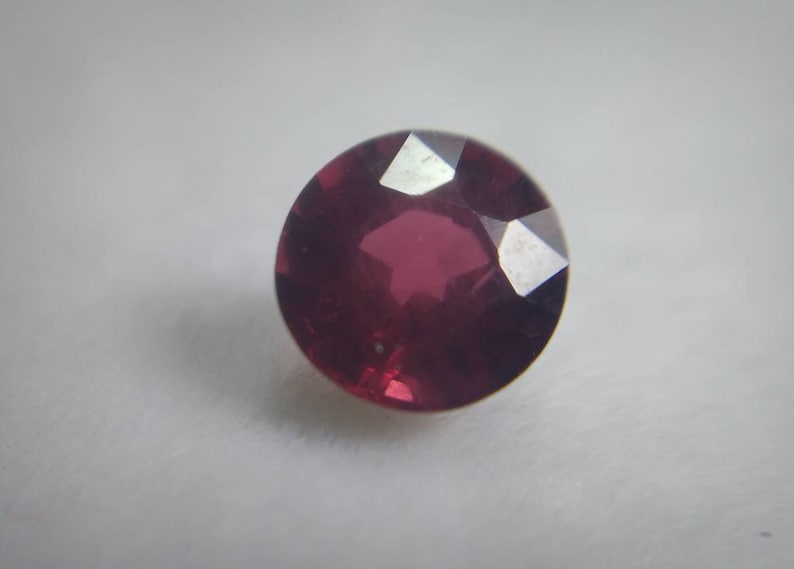 May include: A round, faceted, deep red gemstone. The gem has a polished surface with multiple facets that reflect light. The stone is set against a plain, light background, highlighting its colour and shape. This gemstone could be used in jewellery.