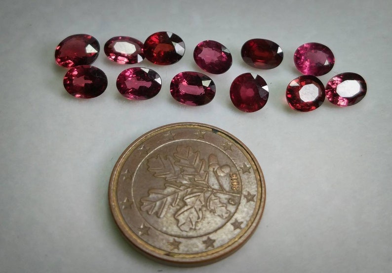 May include: A collection of oval-shaped, deep red gemstones displayed on a white background. A Euro coin is positioned at the bottom for scale. The gemstones exhibit slight variations in colour and clarity.