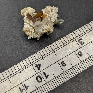 May include: A cluster of crystals featuring a mix of clear, brown, and green hues. The crystals are set on a beige matrix, with a metal ruler providing scale. The composition is natural and mineral-focused.