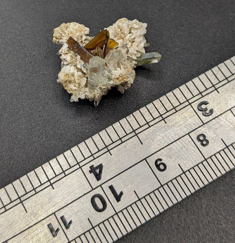 May include: A cluster of crystals featuring a mix of clear, brown, and green hues. The crystals are set on a beige matrix, with a metal ruler providing scale. The composition is natural and mineral-focused.