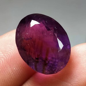 Könnte beinhalten: Ein facettierter, oval geformter Edelstein mit tiefvioletter Farbe, die im Zentrum in ein helleres, rötliches Violett übergeht. Die polierten Oberflächen des Steins reflektieren das Licht und zeigen seine innere Struktur und Klarheit. Geeignet für Schmuck.
