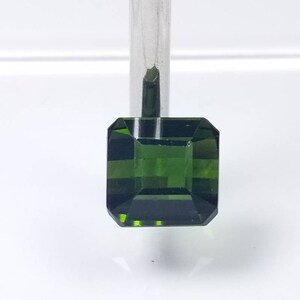 Pode incluir: Uma pedra preciosa verde esmeralda facetada com um corte quadrado. A gema &eacute; segurada por um metal de cor prateada. As facetas da gema refletem a luz, criando um efeito brilhante. O fundo &eacute; uma superf&iacute;cie branca lisa.