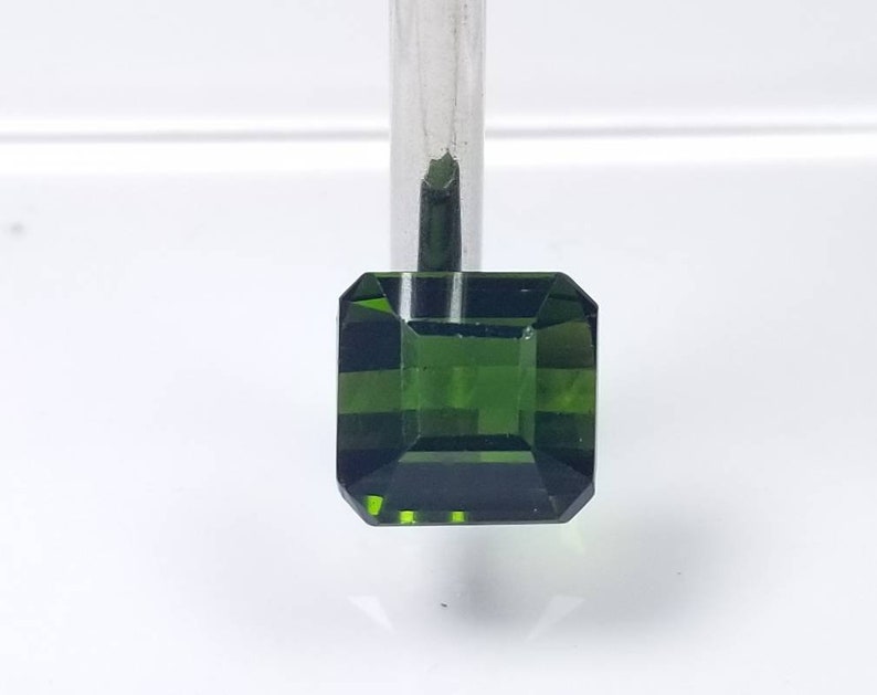 Pode incluir: Uma pedra preciosa verde esmeralda facetada com um corte quadrado. A gema &eacute; segurada por um metal de cor prateada. As facetas da gema refletem a luz, criando um efeito brilhante. O fundo &eacute; uma superf&iacute;cie branca lisa.