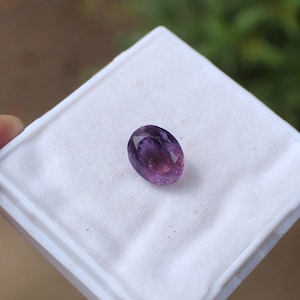 May include: An oval-cut, faceted amethyst gemstone. The gem is a deep purple color with lighter purple and pink hues. It is set on a white surface, with a blurred green background.