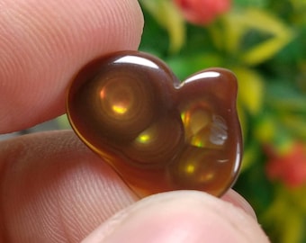 May include: A polished, heart-shaped gemstone with a rich, brown color and swirling patterns. The stone has a glossy surface with iridescent flashes of yellow and green. The gemstone is held between fingers, showcasing its unique shape and color.