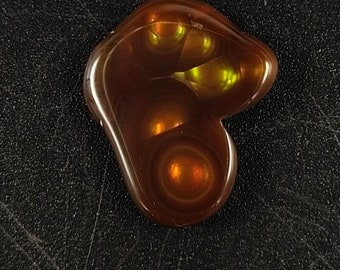 May include: A polished, cabochon-cut gemstone with a rich, warm brown color and internal patterns. The stone has a smooth, rounded shape with areas of orange and yellow light refraction.