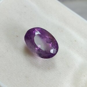 May include: A faceted, oval-shaped amethyst gemstone. The gem is a deep purple color with lighter, translucent areas. The cut of the stone creates multiple reflective surfaces, enhancing its brilliance. The gemstone is set against a white background.
