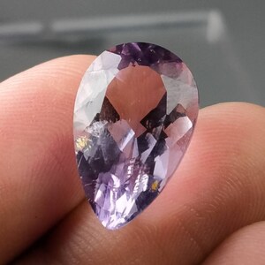 Puede incluir: Una gema facetada en forma de pera con tonos morados. La gema tiene una superficie pulida con m&uacute;ltiples facetas que reflejan la luz. La piedra se sostiene entre los dedos, mostrando su tama&ntilde;o y forma. La gema parece ser un tipo de amatista.