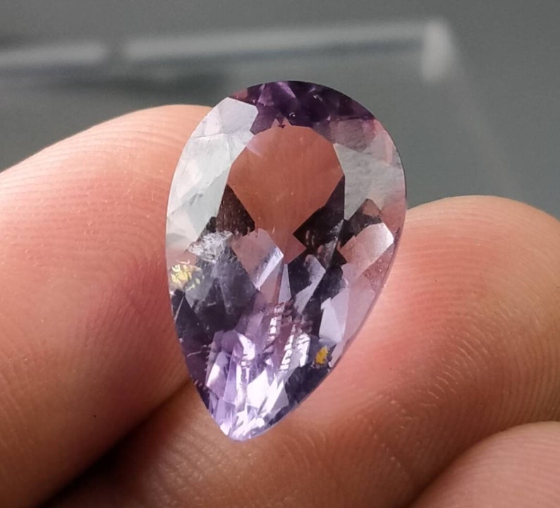 Puede incluir: Una gema facetada en forma de pera con tonos morados. La gema tiene una superficie pulida con m&uacute;ltiples facetas que reflejan la luz. La piedra se sostiene entre los dedos, mostrando su tama&ntilde;o y forma. La gema parece ser un tipo de amatista.