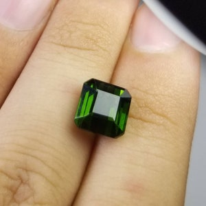 Pode incluir: Uma pedra preciosa verde esmeralda facetada com formato retangular. A gema &eacute; segurada em uma m&atilde;o, mostrando sua clareza e cor profunda. O corte da pedra permite que a luz se reflita.