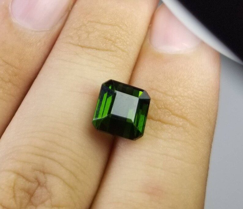 Pode incluir: Uma pedra preciosa verde esmeralda facetada com formato retangular. A gema &eacute; segurada em uma m&atilde;o, mostrando sua clareza e cor profunda. O corte da pedra permite que a luz se reflita.