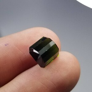 Pode incluir: Uma pedra preciosa verde-escura facetada com formato retangular. A superf&iacute;cie polida da gema possui m&uacute;ltiplas facetas que refletem a luz. A cor transita de um verde escuro, quase preto, para um tom verde-amarelado mais claro.