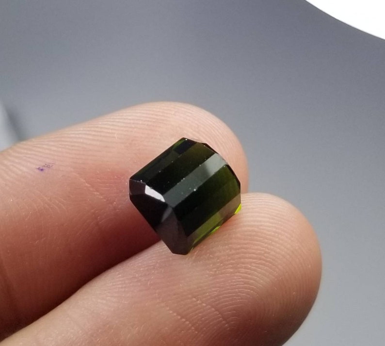 Pode incluir: Uma pedra preciosa verde-escura facetada com formato retangular. A superf&iacute;cie polida da gema possui m&uacute;ltiplas facetas que refletem a luz. A cor transita de um verde escuro, quase preto, para um tom verde-amarelado mais claro.