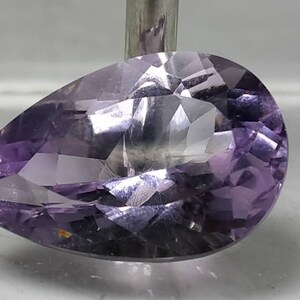 Puede incluir: Una gema facetada en forma de pera, de color p&uacute;rpura claro. La gema est&aacute; tallada con m&uacute;ltiples facetas, creando un efecto brillante. La piedra est&aacute; sujeta por una varilla met&aacute;lica, mostrando su forma y color.