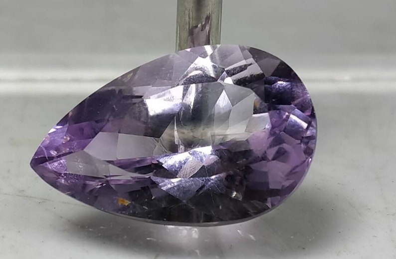 Puede incluir: Una gema facetada en forma de pera, de color p&uacute;rpura claro. La gema est&aacute; tallada con m&uacute;ltiples facetas, creando un efecto brillante. La piedra est&aacute; sujeta por una varilla met&aacute;lica, mostrando su forma y color.