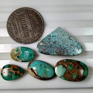 Puede incluir: Una colecci&oacute;n de piedras preciosas de turquesa en varias formas, incluyendo ovaladas y en forma de l&aacute;grima, con patrones de matriz marr&oacute;n. Se incluye una moneda de un centavo estadounidense para referencia de tama&ntilde;o. Las piedras est&aacute;n sobre una superficie blanca.